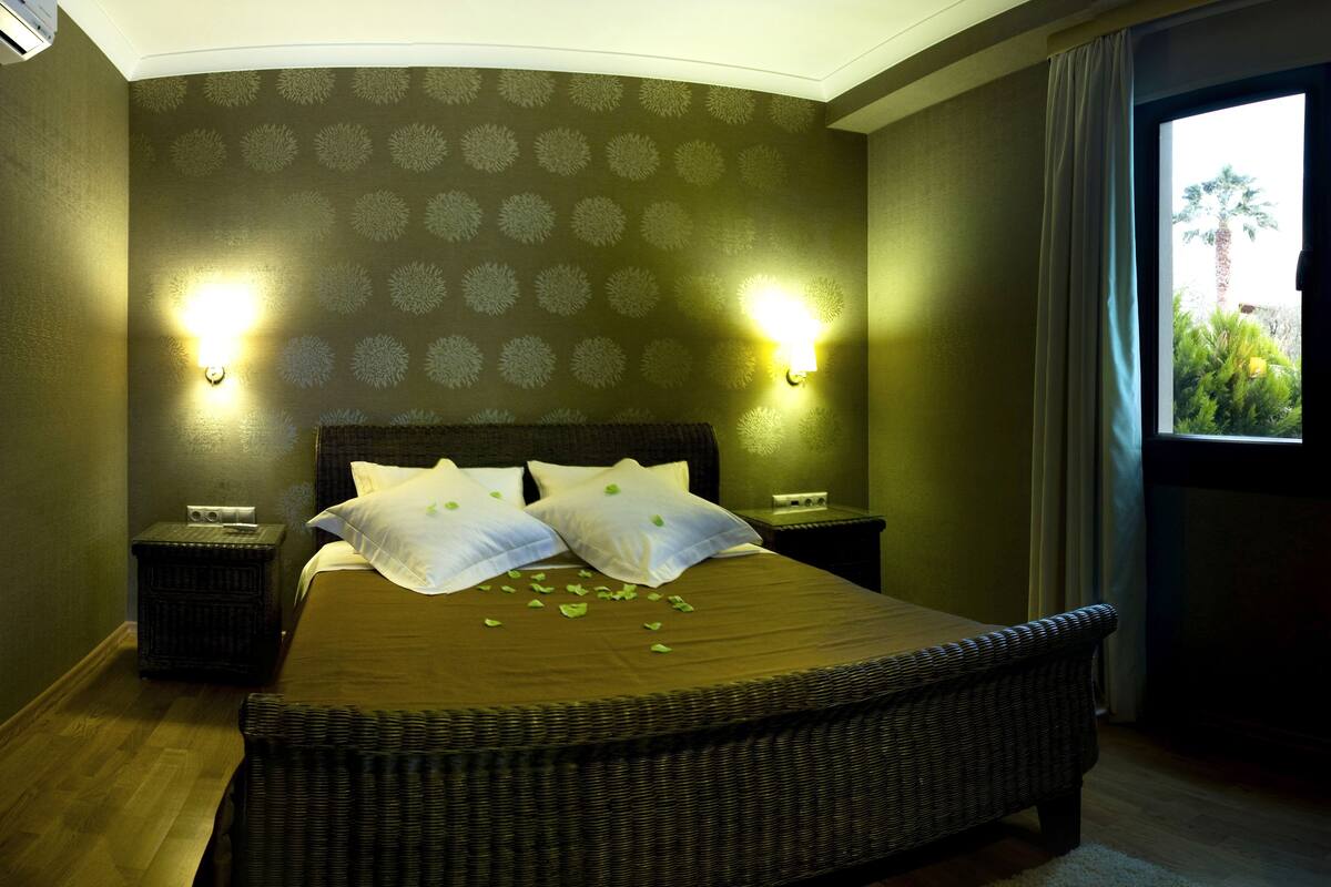 Habitación de hotel con una cama grande, dos mesitas de noche, una mesilla auxiliar, una silla de mimbre y una ventana con vistas a la vegetación.
