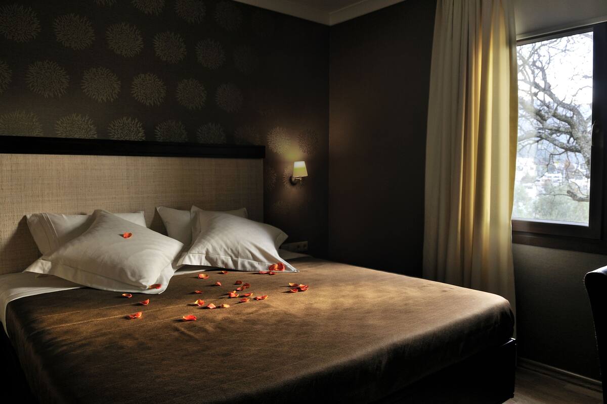 Una habitación de hotel con una cama grande, una mesita de noche, una lámpara y una ventana con cortinas.