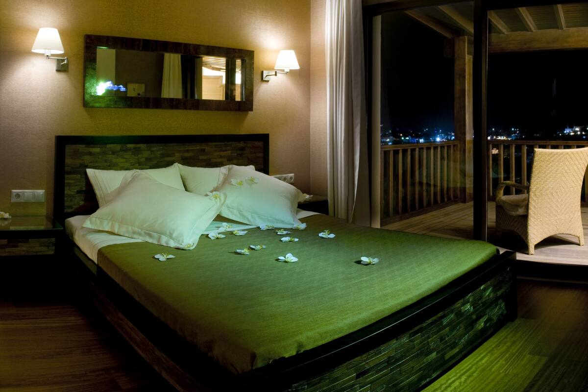 Una habitación de hotel con cama, mesita de noche, espejo y balcón con una silla.