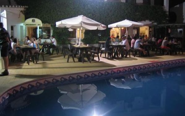 Ein Außenbereich zum Essen mit Tischen, Stühlen, Sonnenschirmen und einem Pool im Vordergrund.