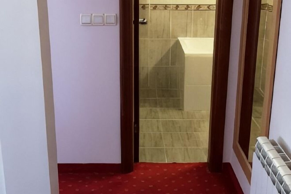 A narrow hallway with a red carpet, a door leading to a bathroom with a tiled wall, and a radiator on the right.