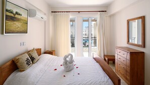A neatly made bed with a white comforter and a swan-shaped towel centerpiece, a wooden dresser, a painting on the wall, and a balcony with curtains. - St Nicolas Elegant Residence (Paphos)