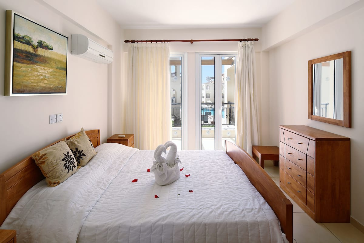 A neatly made bed with a white comforter and a swan-shaped towel centerpiece, a wooden dresser, a painting on the wall, and a balcony with curtains.