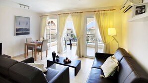 A modern living room with a sofa, a coffee table, and a dining table with chairs. There is a balcony with yellow curtains and a view of the outside. - St Nicolas Elegant Residence (Paphos)