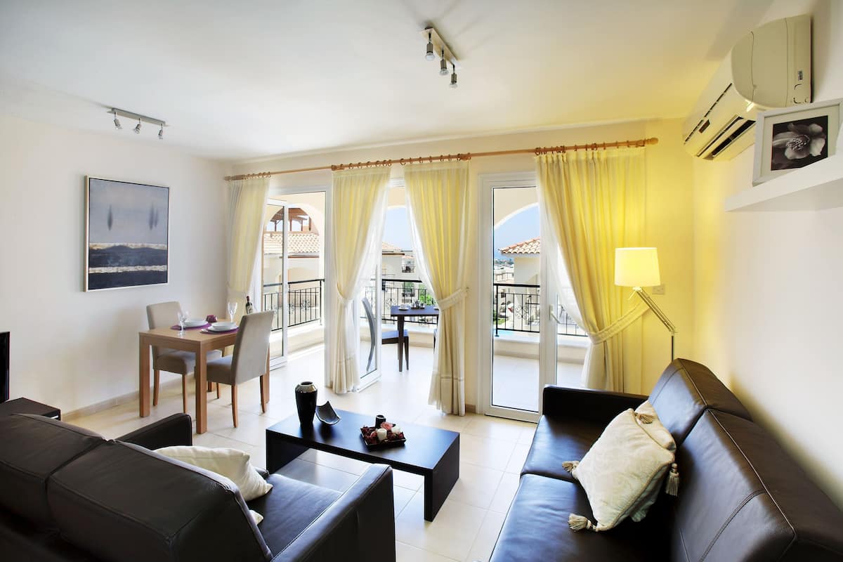 A modern living room with a sofa, a coffee table, and a dining table with chairs. There is a balcony with yellow curtains and a view of the outside.