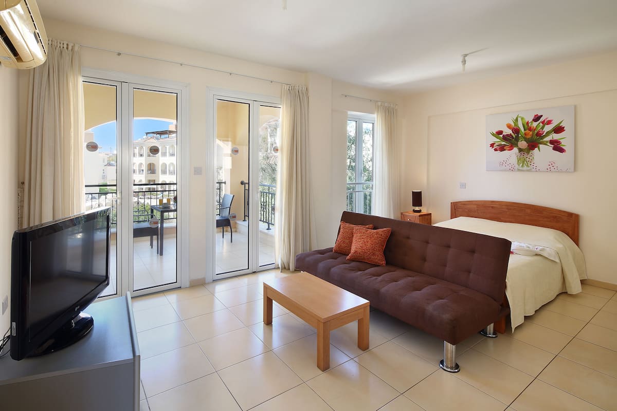 A hotel room with a bed, a sofa, a TV, a coffee table, and a balcony with a view of buildings.