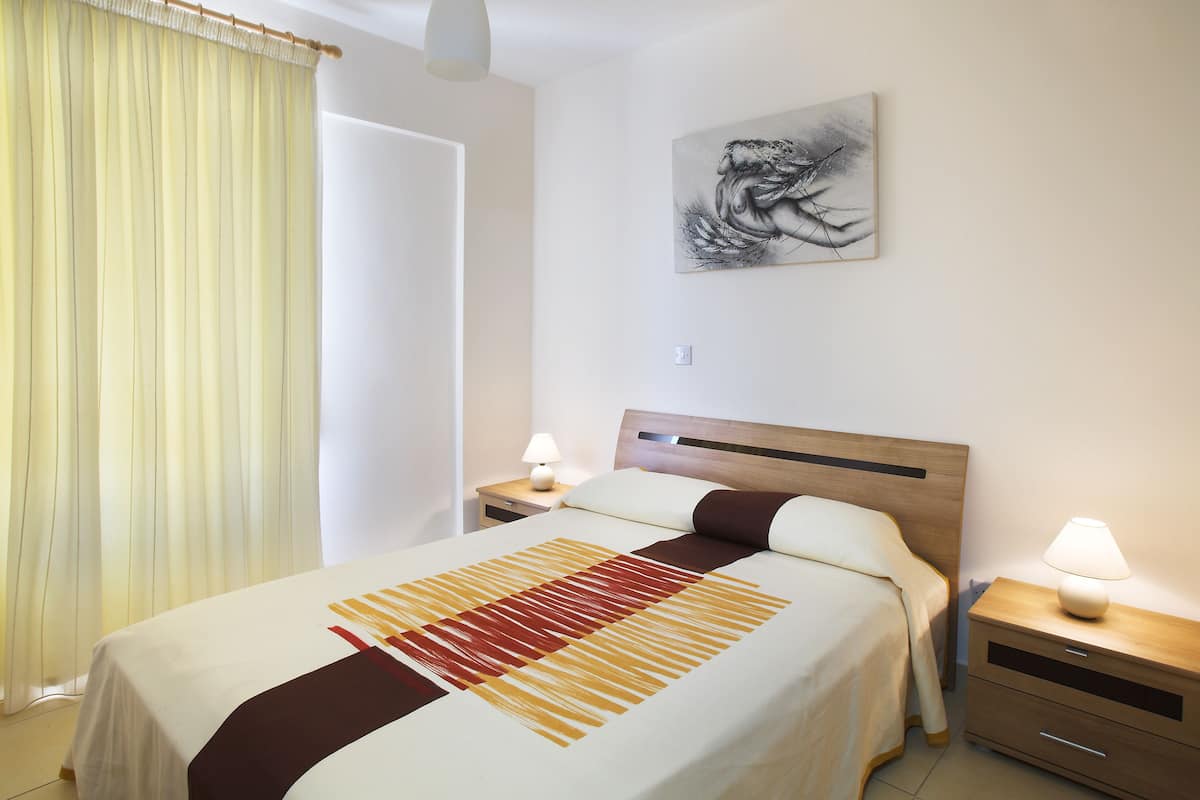 A hotel room with a bed, a nightstand with a lamp, a painting on the wall, and a window with curtains.