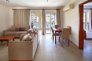 A modern living room with a sofa, coffee table, dining table, and chairs. There is a large window with curtains, an air conditioning unit, and a view of buildings and a pool. - St Nicolas Elegant Residence (Paphos)