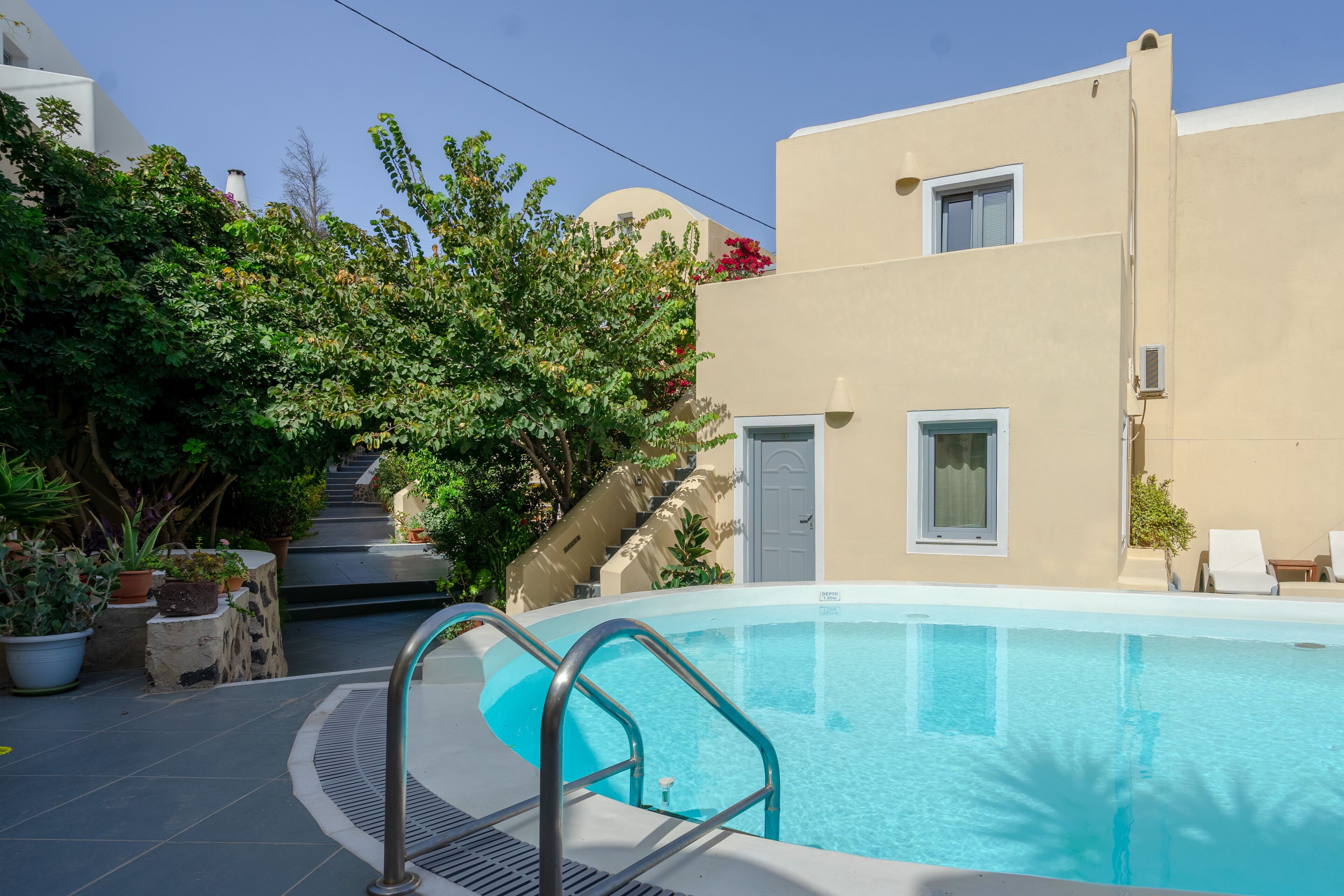 A swimming pool with a metal ladder, surrounded by potted plants and a building with a beige facade and a door.