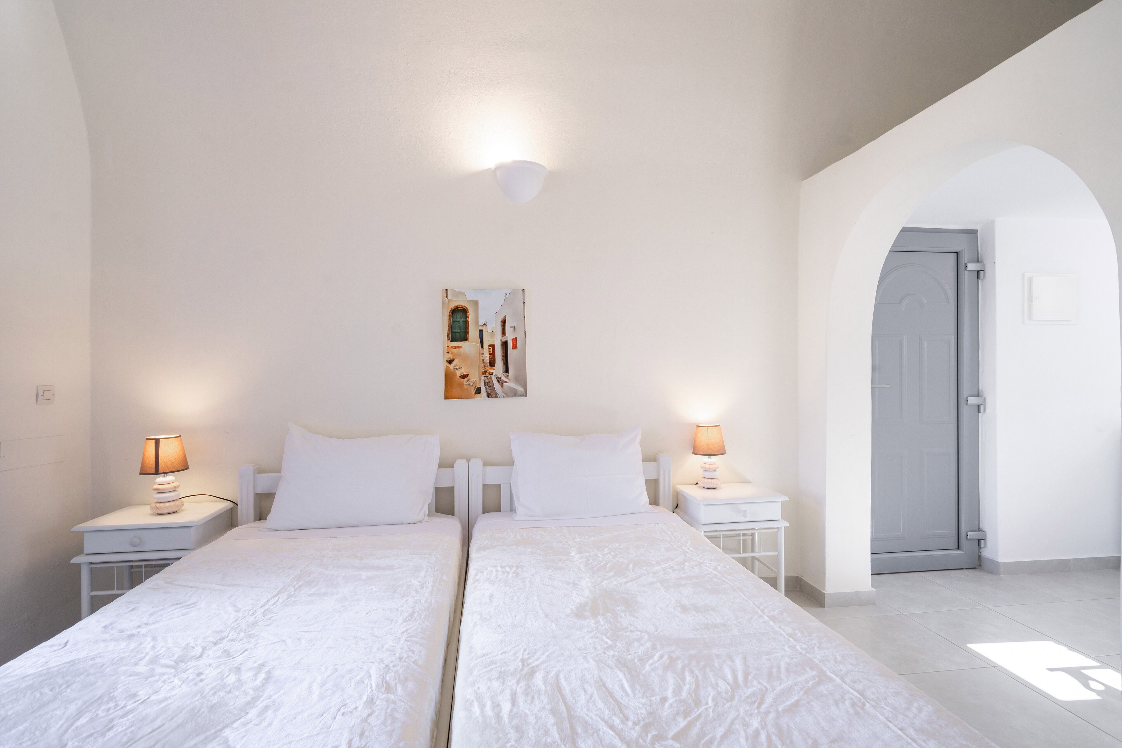 A double bed with white linens, two bedside tables with lamps, a wall-mounted picture, and a door with a grey frame.