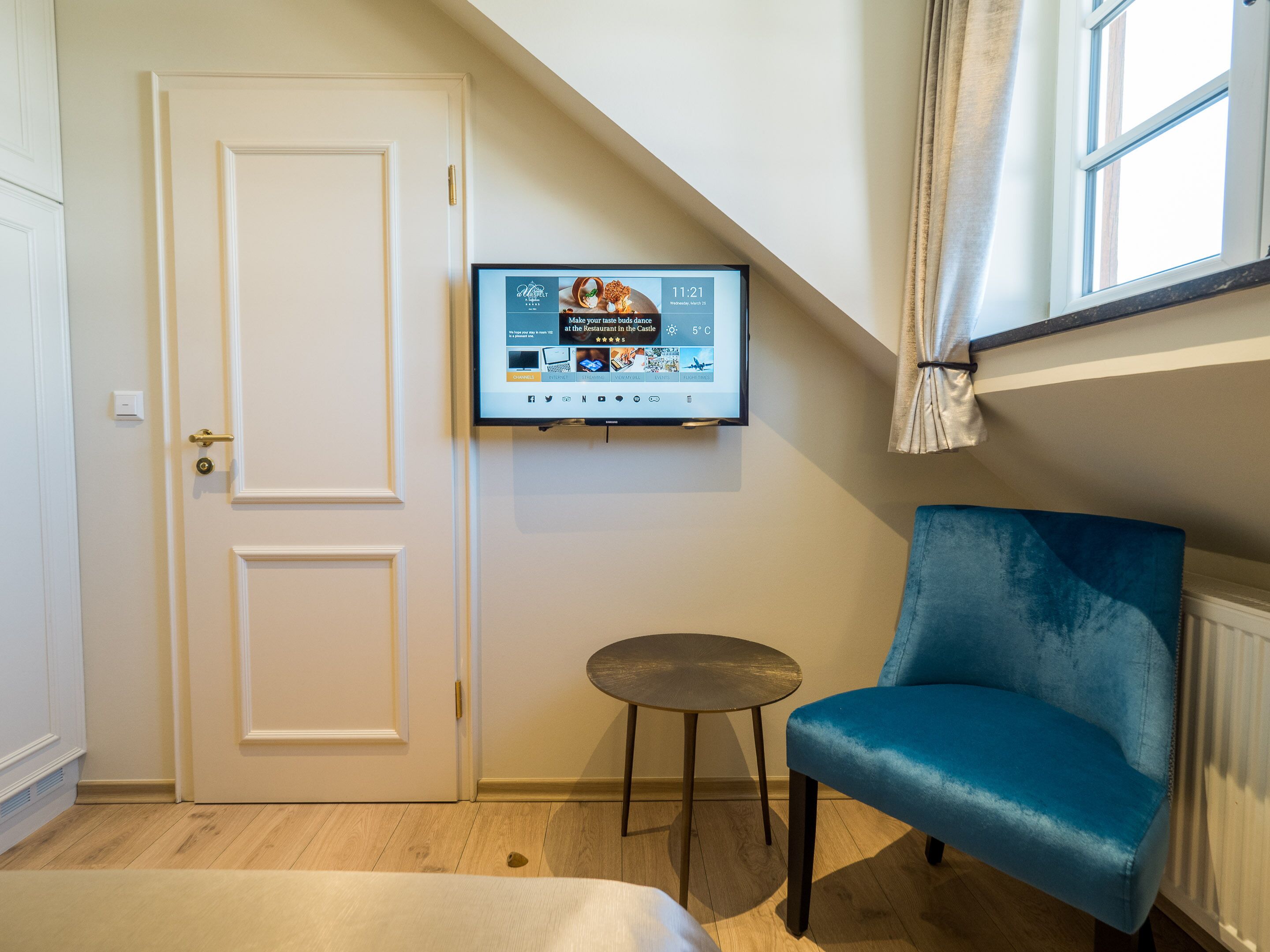 Una habitació amb una porta blanca, un televisor fixat amb un menú de restaurant i una taula rodona petita, juntament amb una cadira blava.