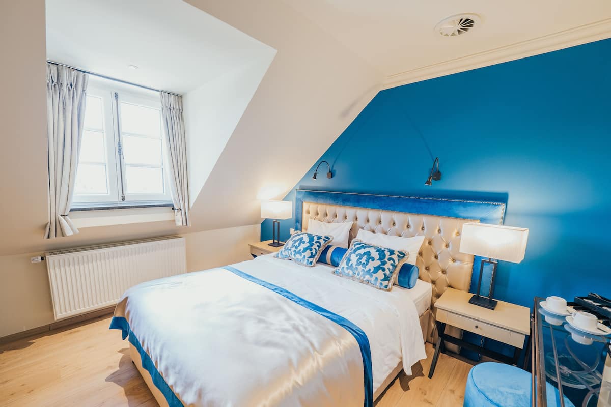 A bedroom with a sloped ceiling, a bed with a white and blue bedspread, two bedside lamps, a nightstand, and a blue ottoman.