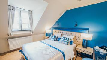 A bedroom with a sloped ceiling, a bed with a white and blue bedspread, two bedside lamps, a nightstand, and a blue ottoman.