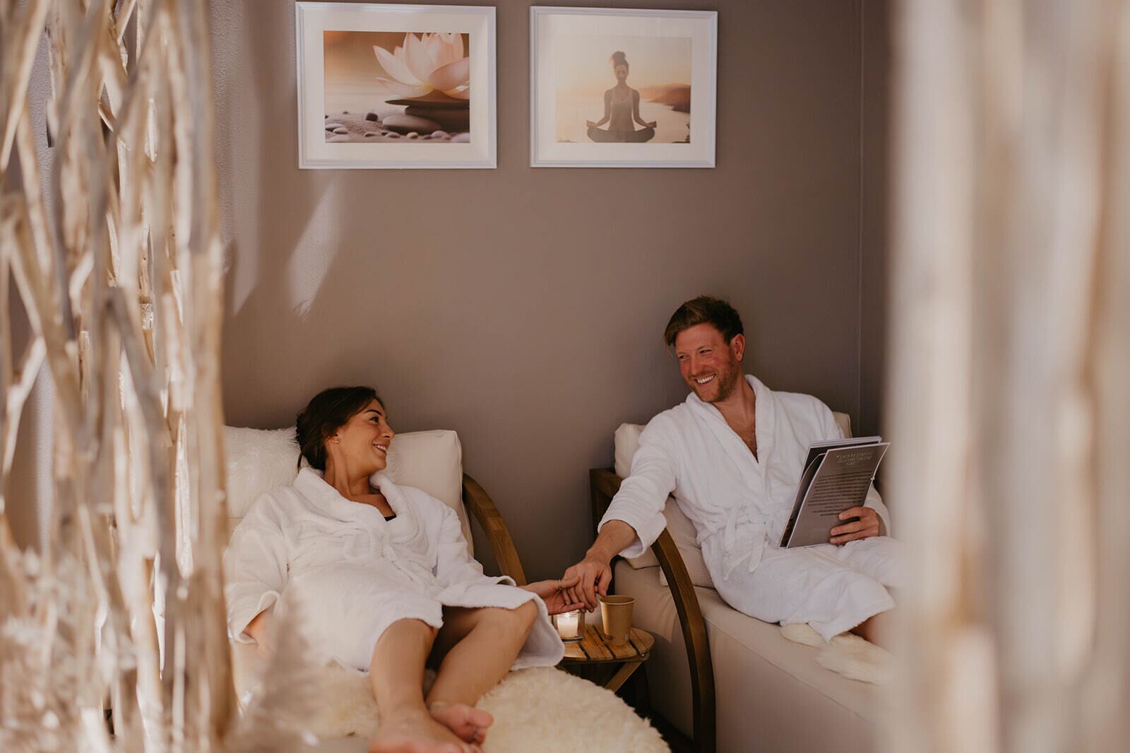 A spa room with two individuals in white robes, a wooden table with a cup, and framed pictures on the wall.