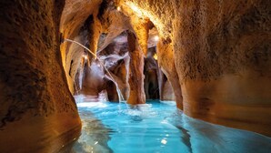 Une grotte ornée de stalactites et de stalagmites, avec une mare d’eau au centre.