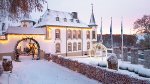 A snow-covered building with a large archway entrance, a spherical light decoration, and a flagpole. - Chateau d'Urspelt (Clervaux)