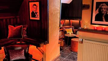 Un espacio de bar acogedor con una banqueta de cuero, una barra de madera y piso de baldosas. Hay dos mesitas redondas con velas y menús.