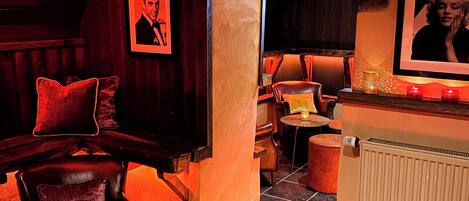 Un coin bar accueillant avec un banquette en cuir, un comptoir en bois et un sol carrelé. On y trouve deux petites tables rondes, décorées de bougies et accompagnées de menus.