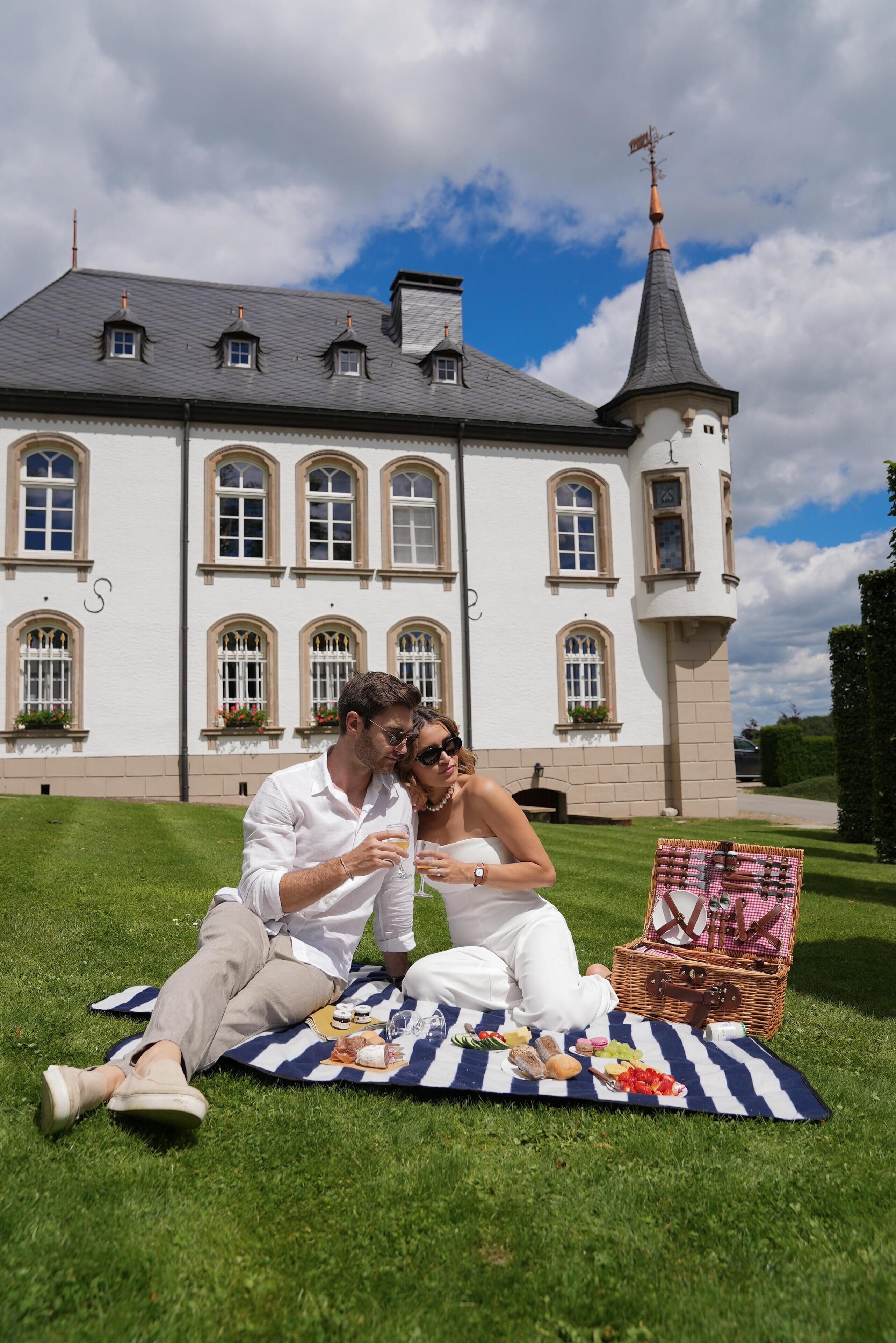 A couple sitting on a picnic blanket with a picnic basket in front of a large white building with multiple windows and a tower.
