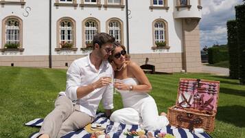 A couple sitting on a picnic blanket with a picnic basket in front of a large white building with multiple windows and a tower.