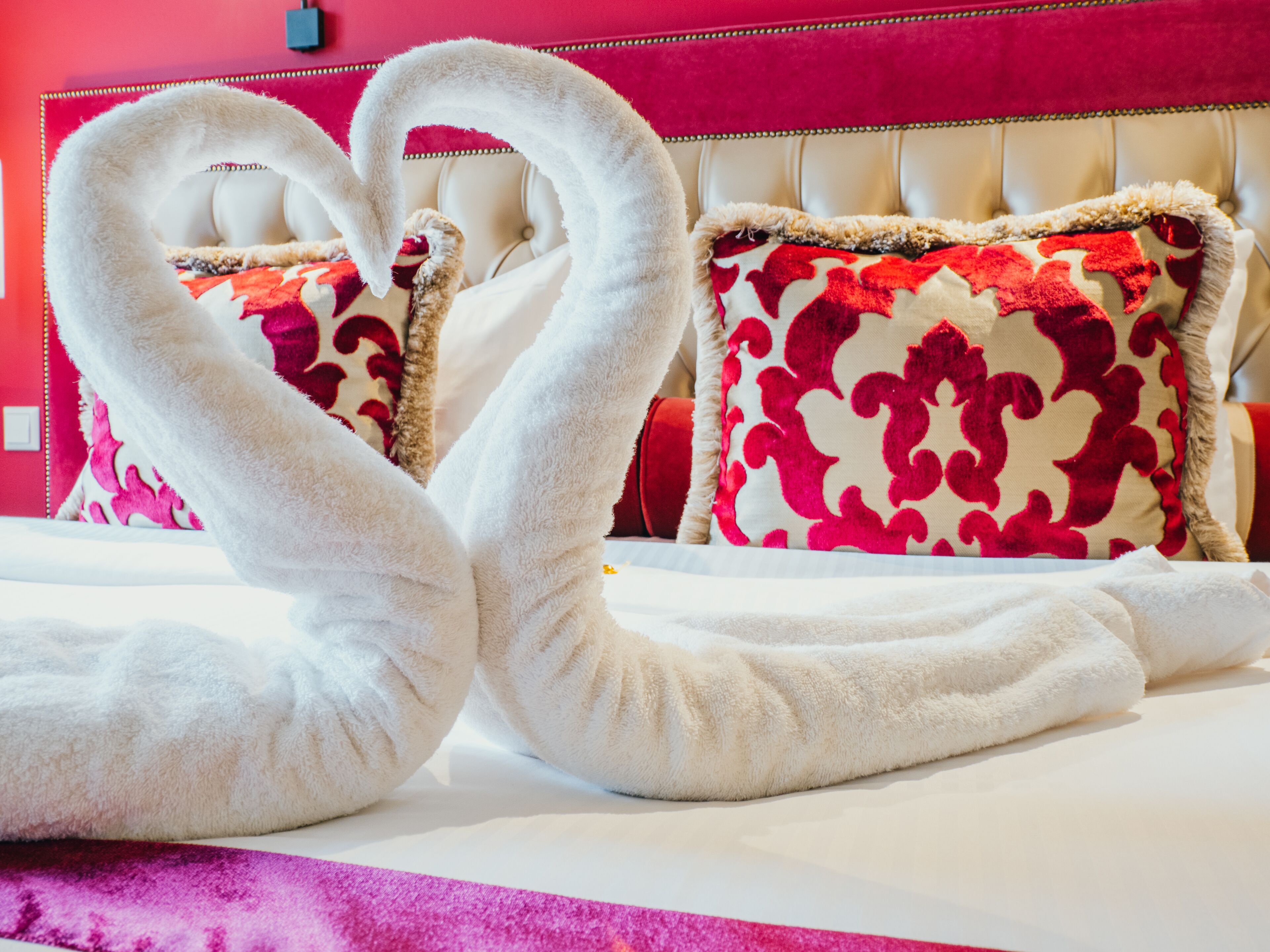 A bed with white towels arranged in a heart shape, red and white patterned pillows, and a tufted headboard.