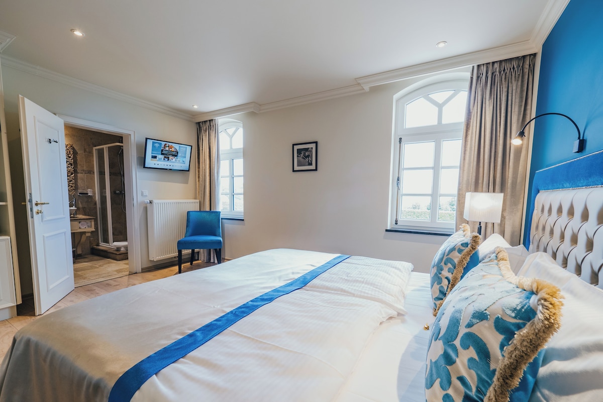 Habitación de hotel con una cama grande, televisión, ventana con cortinas y baño visible a través de una puerta abierta.