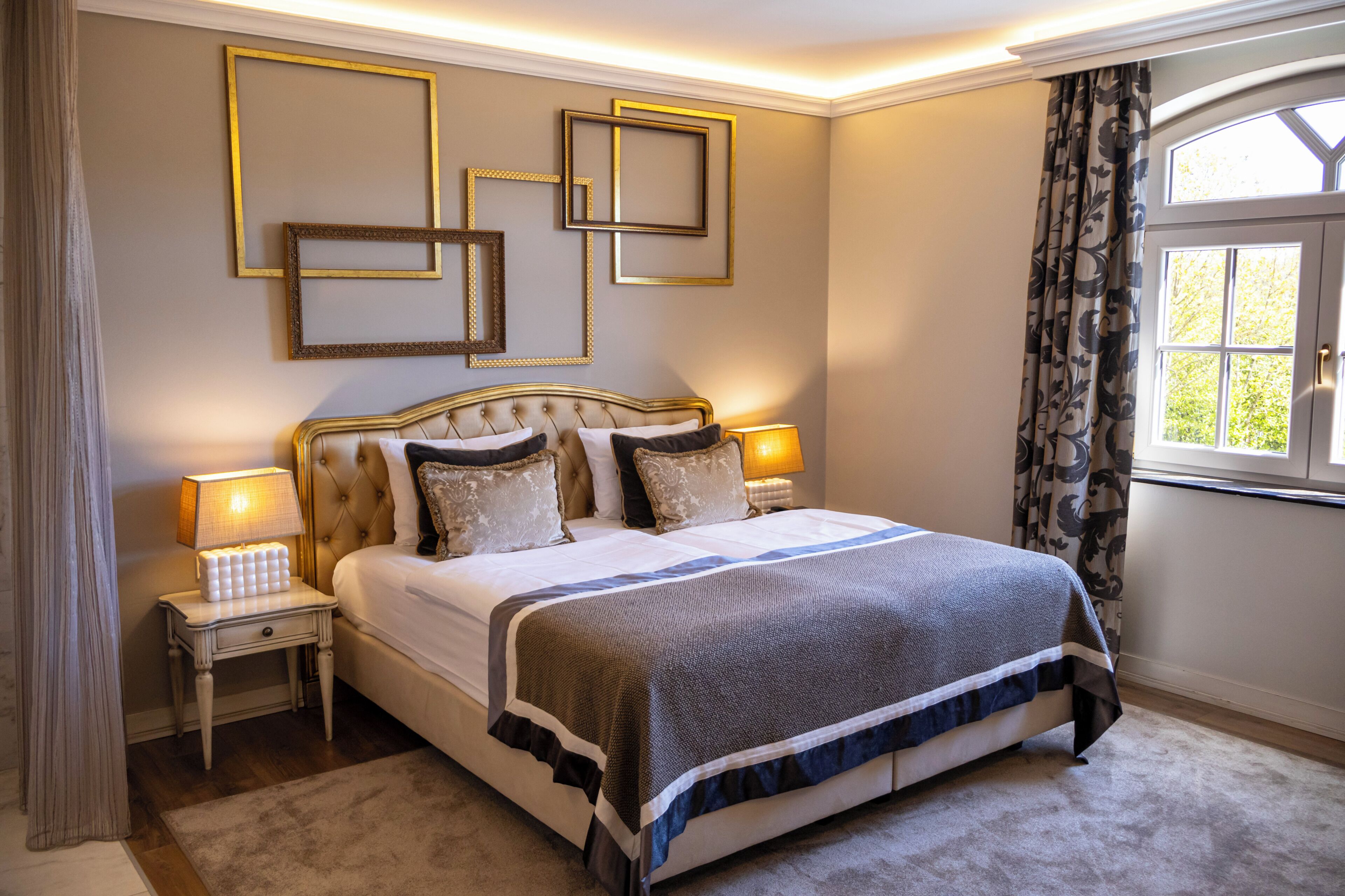 Un dormitorio con una cama grande, mesitas de noche, marcos de cuadros en la pared y una ventana con cortinas.