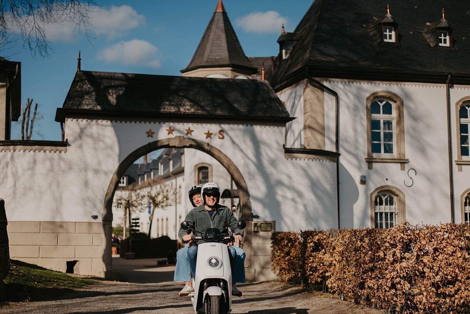 A couple on a scooter approaching a building with a large archway and stars above it.
