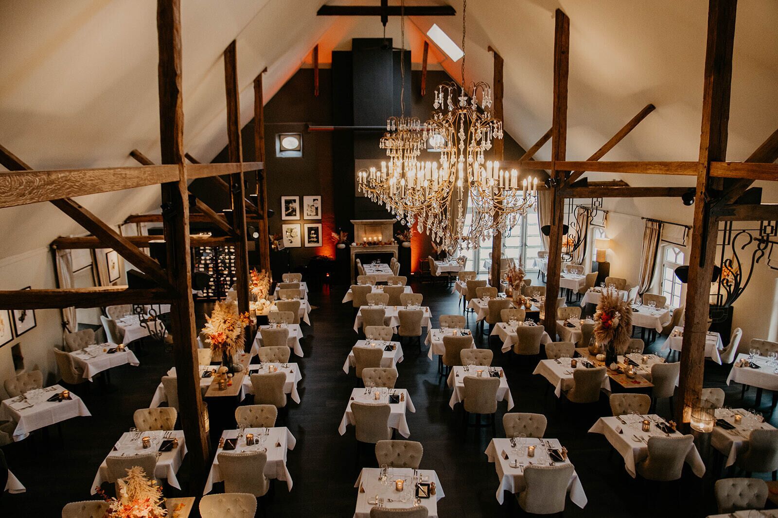 A large, elegantly decorated hall with numerous tables and chairs, high ceilings, and a grand chandelier.