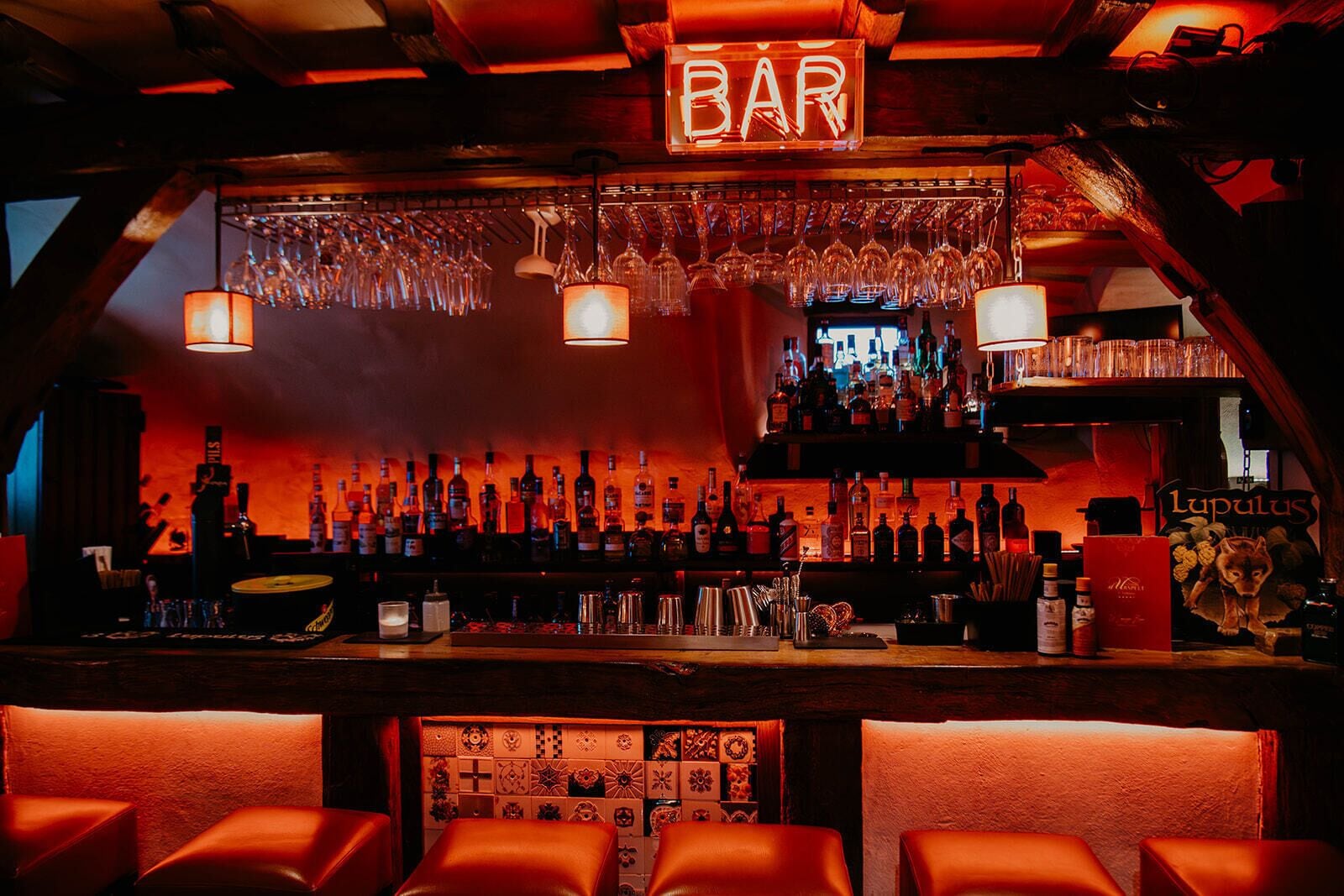 A bar area with a neon 'BAR' sign, shelves of liquor bottles, hanging glasses, and leather bar stools.