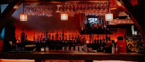 A bar area with a neon 'BAR' sign, shelves of liquor bottles, hanging glasses, and leather bar stools.