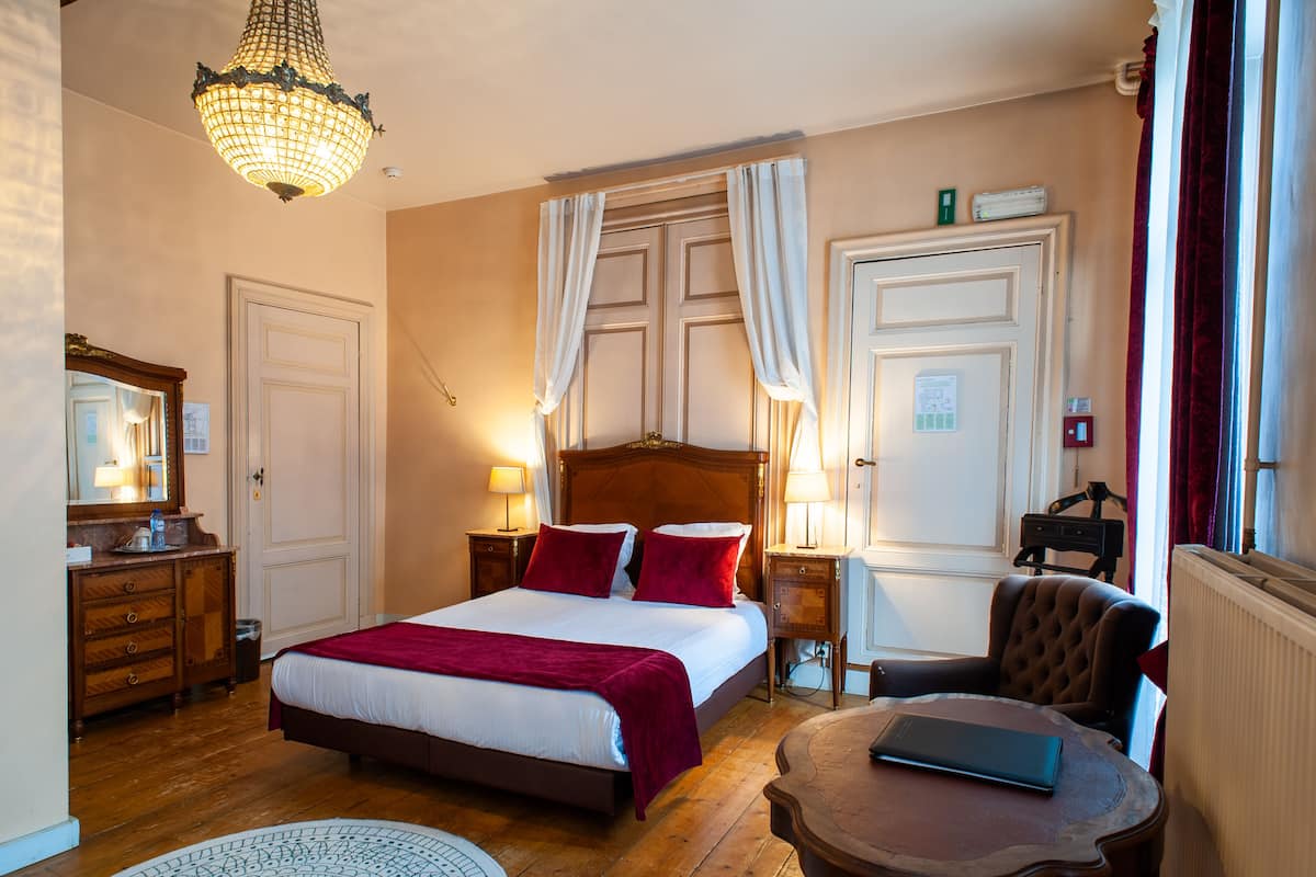 Une chambre d’hôtel avec un lit en bois, une table basse ronde, une chaise, une commode et un lustre.