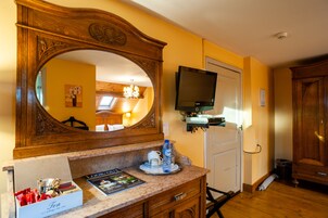 Habitación de hotel con tocador de madera, un espejo grande, un televisor montado en la pared, una mesita con una botella de agua y una taza, y una puerta que da a otra habitación.