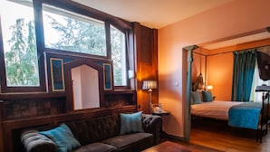 Una habitación con sofá de madera, una mesita, una cama con ropa de cama azul y una ventana con vistas a árboles.