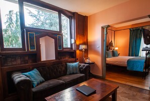 Una habitación con sofá de madera, una mesita, una cama con ropa de cama azul y una ventana con vistas a árboles.