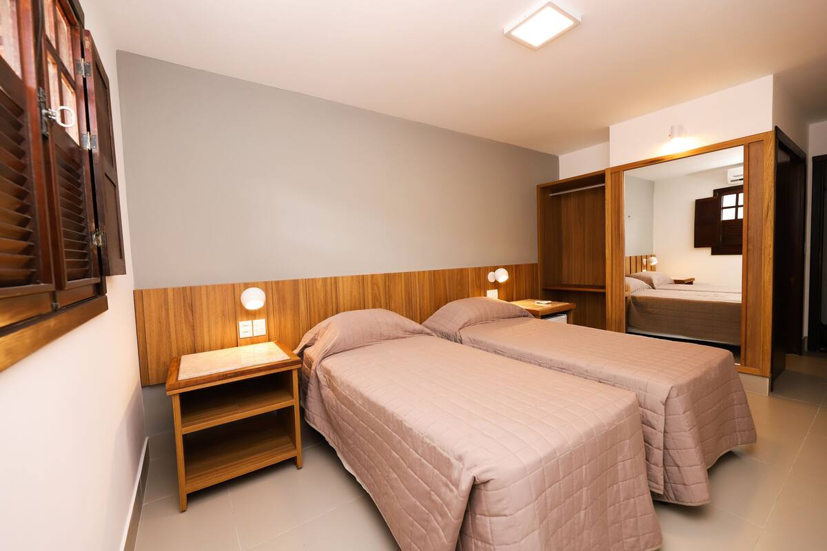 Quarto com duas camas de solteiro, cabeceiras de madeira, mesas de cabeceira com luminárias e um espelho grande.