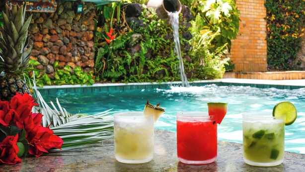 Escena junto a la piscina con un muro de piedra, una fuente de agua y tres bebidas con adornos.