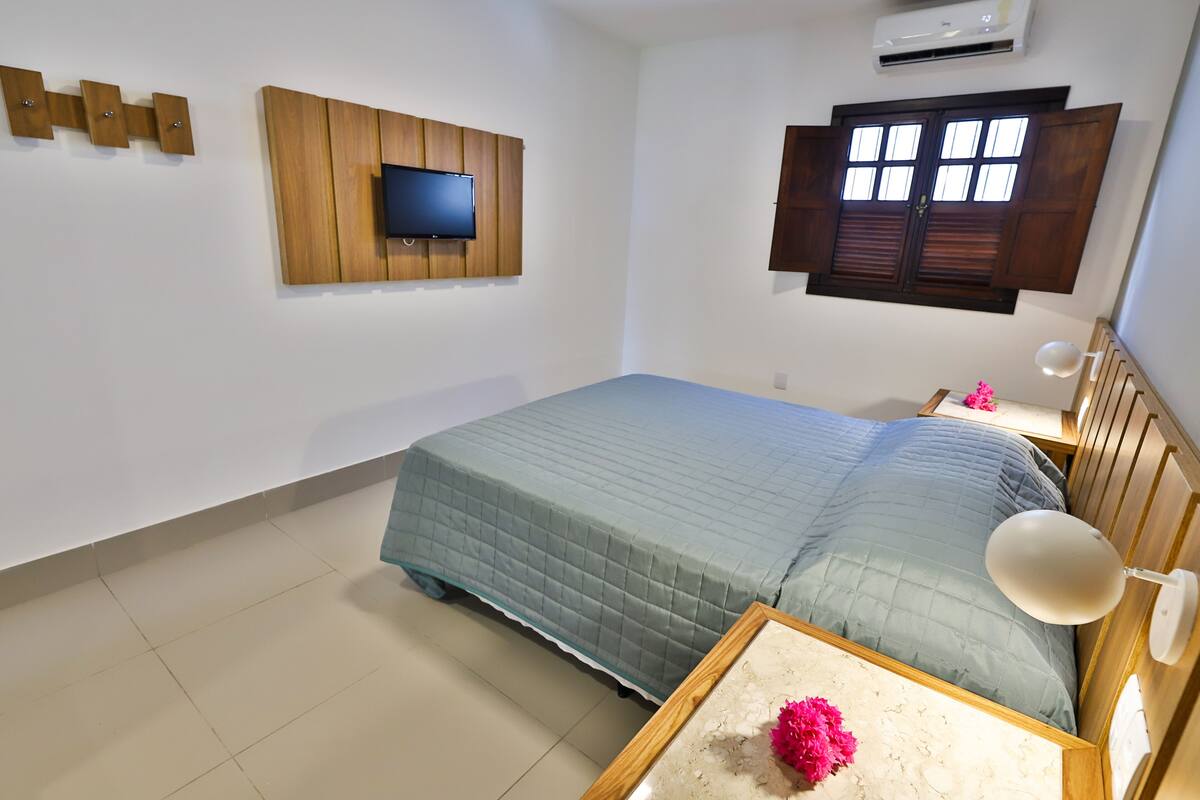 Quarto com cama, televisão fixada num painel de madeira, janela com persianas e mesa de cabeceira com abajur.