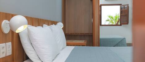 A hotel room with a bed, a bedside lamp, a wooden headboard, a window with shutters, and an air conditioning unit.
