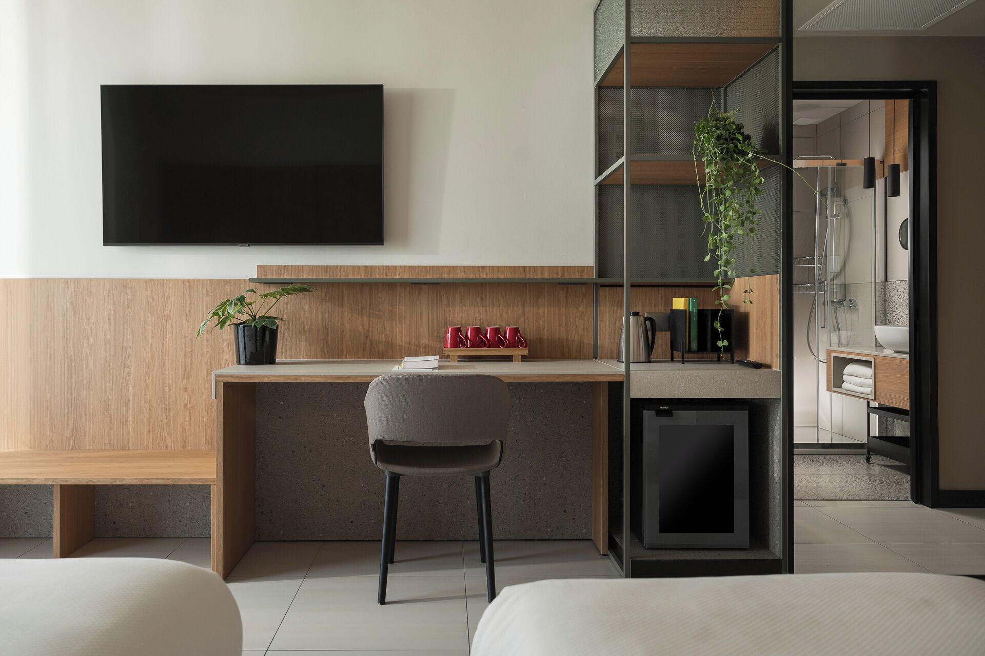 Una habitación de hotel moderna con un escritorio de madera, una silla, un televisor, una planta y un área de ducha.