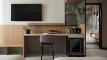 A modern hotel room with a wooden desk, a chair, a TV, a plant, and a shower area.
