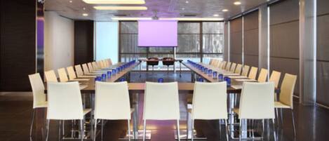 A conference room with a long table, white chairs, and a large screen at the front.