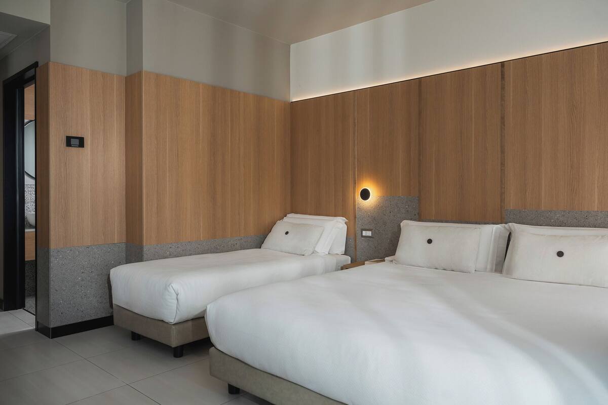 A hotel room with two beds, wooden paneling on the walls, a built-in light, and a small bedside table.