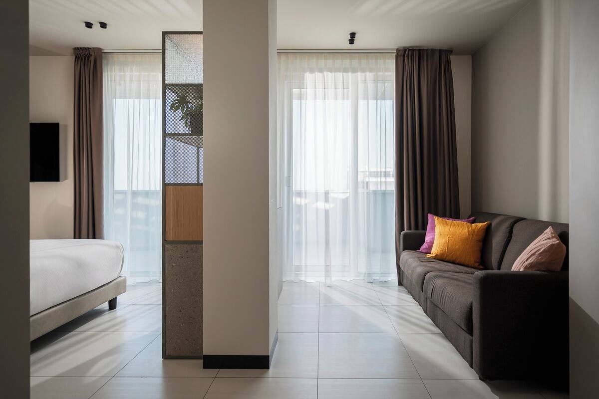 A modern hotel room with a bed, a sofa, and a large window with sheer curtains.