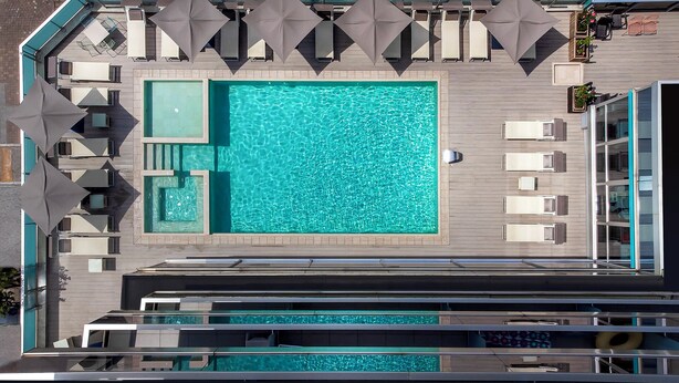 Vista aerea di una zona piscina con lettini e ombrelloni, una vasca idromassaggio separata e una sezione in vetro.