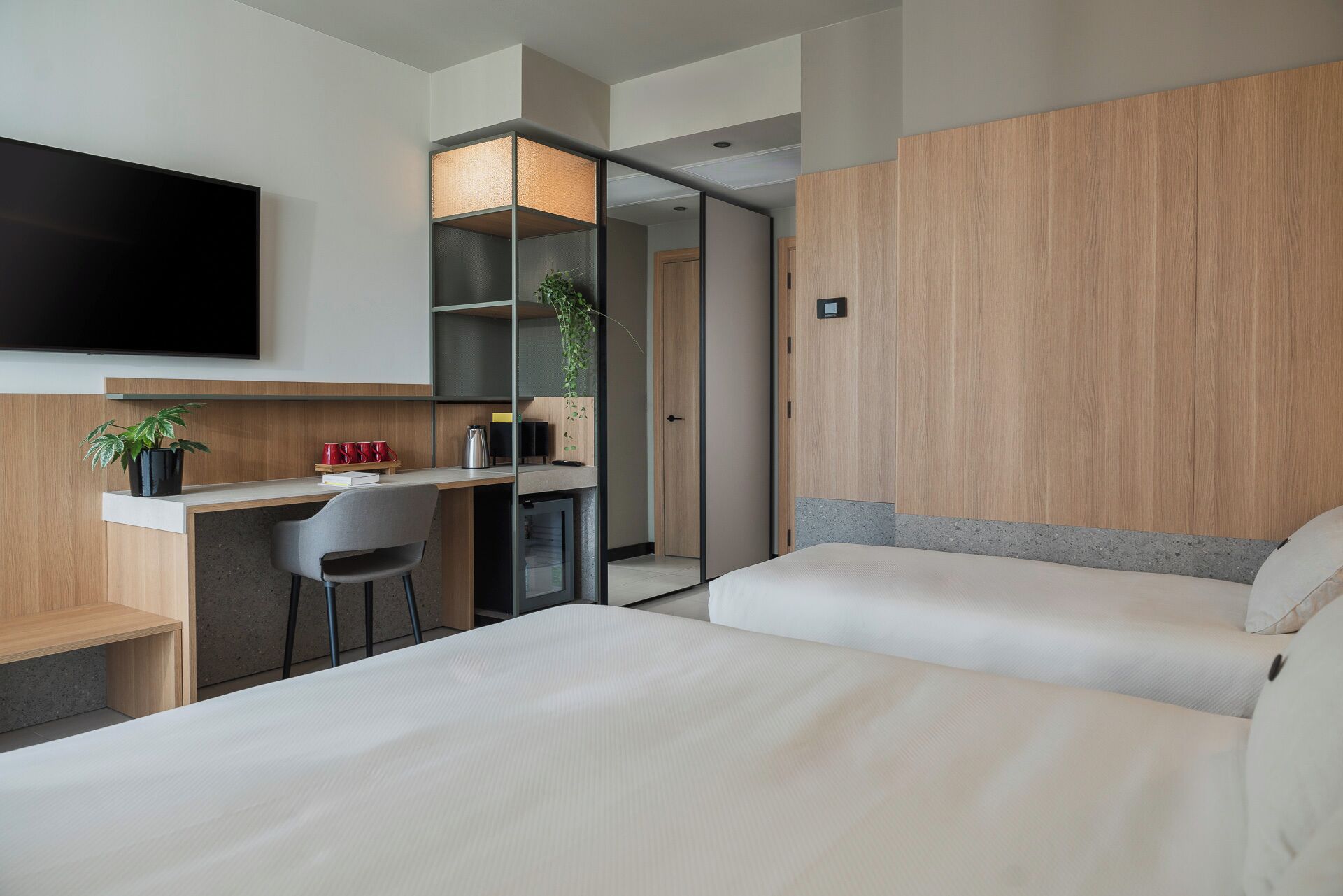 A modern hotel room with a desk, two beds, a TV, and a plant.