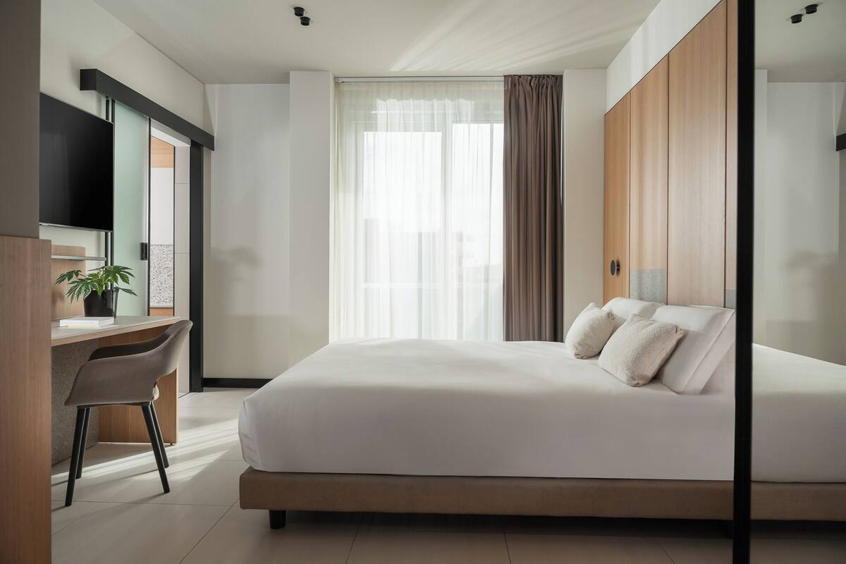 A modern hotel room with a large bed, a desk with a chair, a TV, and a window with curtains.