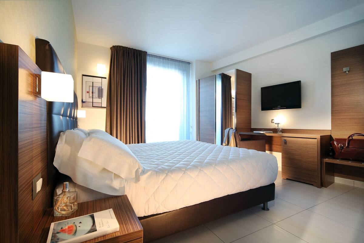 A hotel room with a large bed, a bedside table with a lamp, a TV mounted on the wall, and a wooden nightstand.