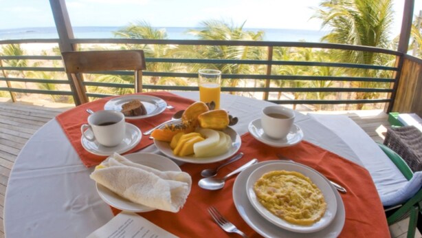 Uma mesa posta com itens de café da manhã, incluindo ovos, torradas e suco, numa varanda com vista para o oceano.