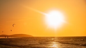 Uma praia com o sol se pondo no oceano, pipas voando e uma terra à distância.